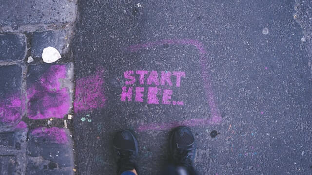 'Start Here' graffiti on road, in front of two feet.